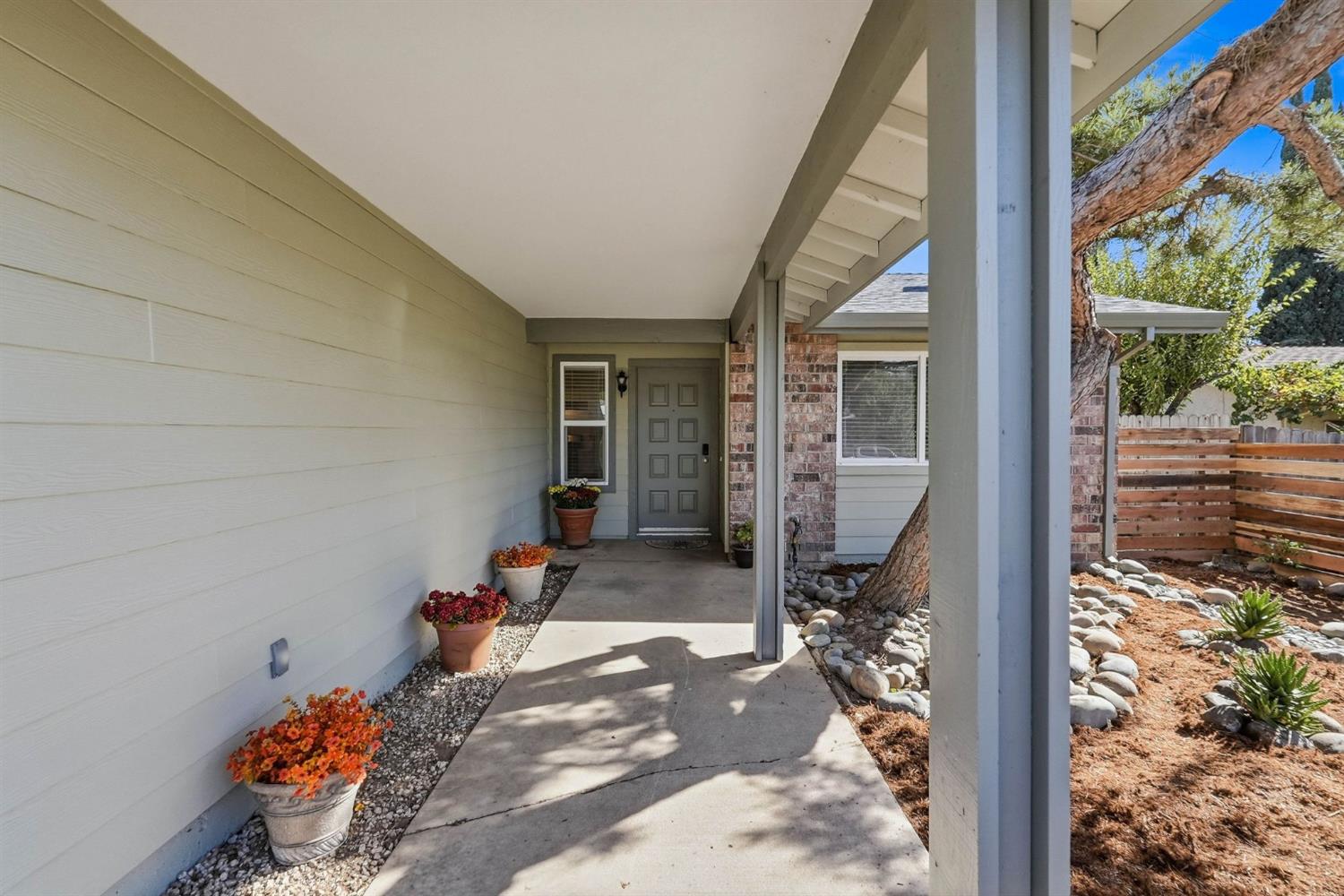 Covered home entrance with a gray paneled door, potted flowers, and a mature tree beside the walkway.