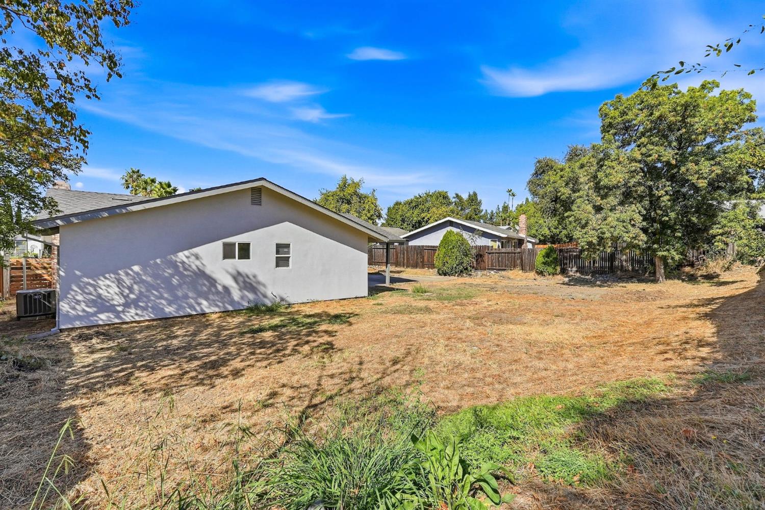 Single-story home beside a broad, dry backyard with mature trees and wooden fencing.