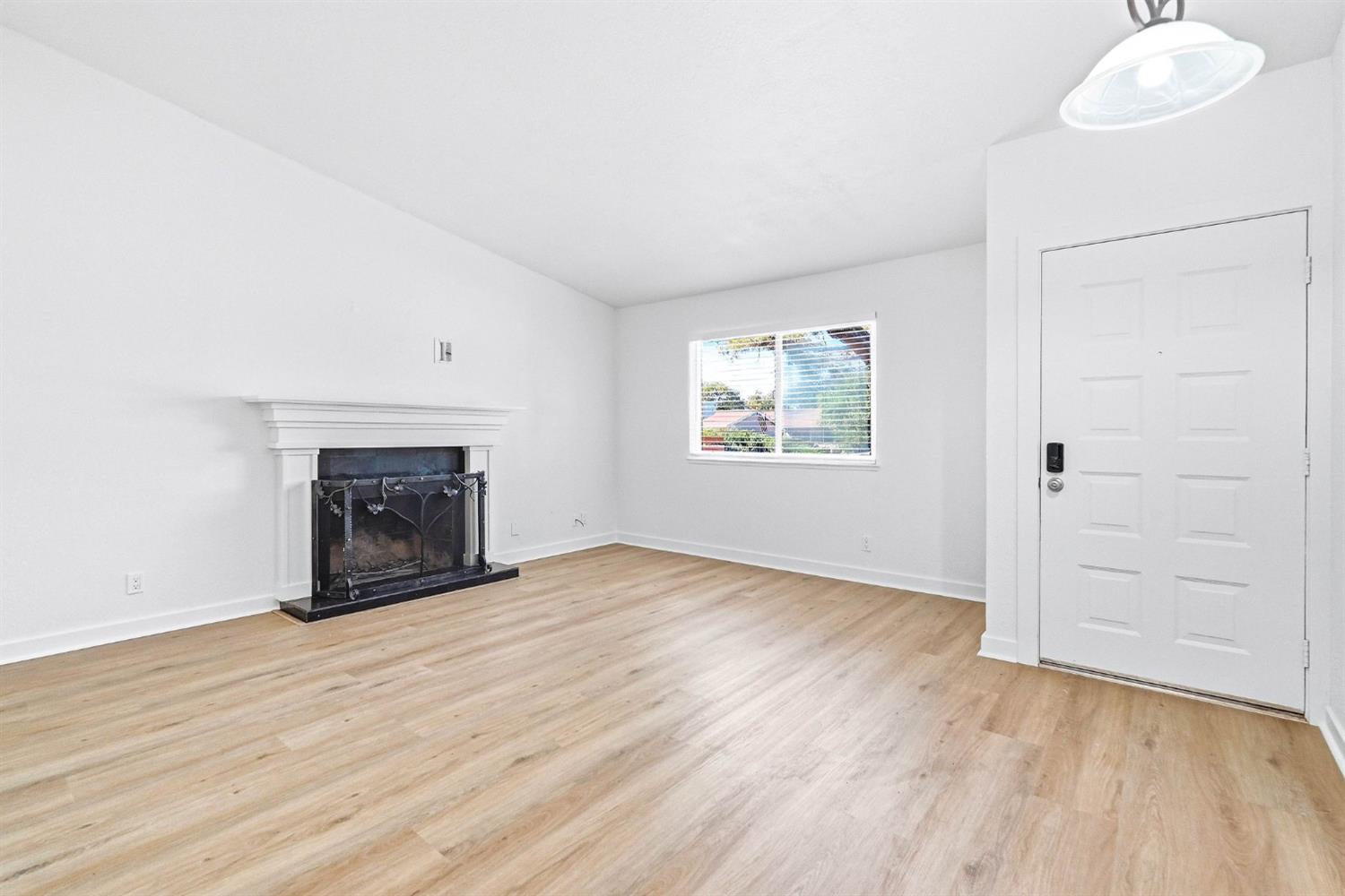 Empty living room with light wood floors, a fireplace, and a window beside the front door.