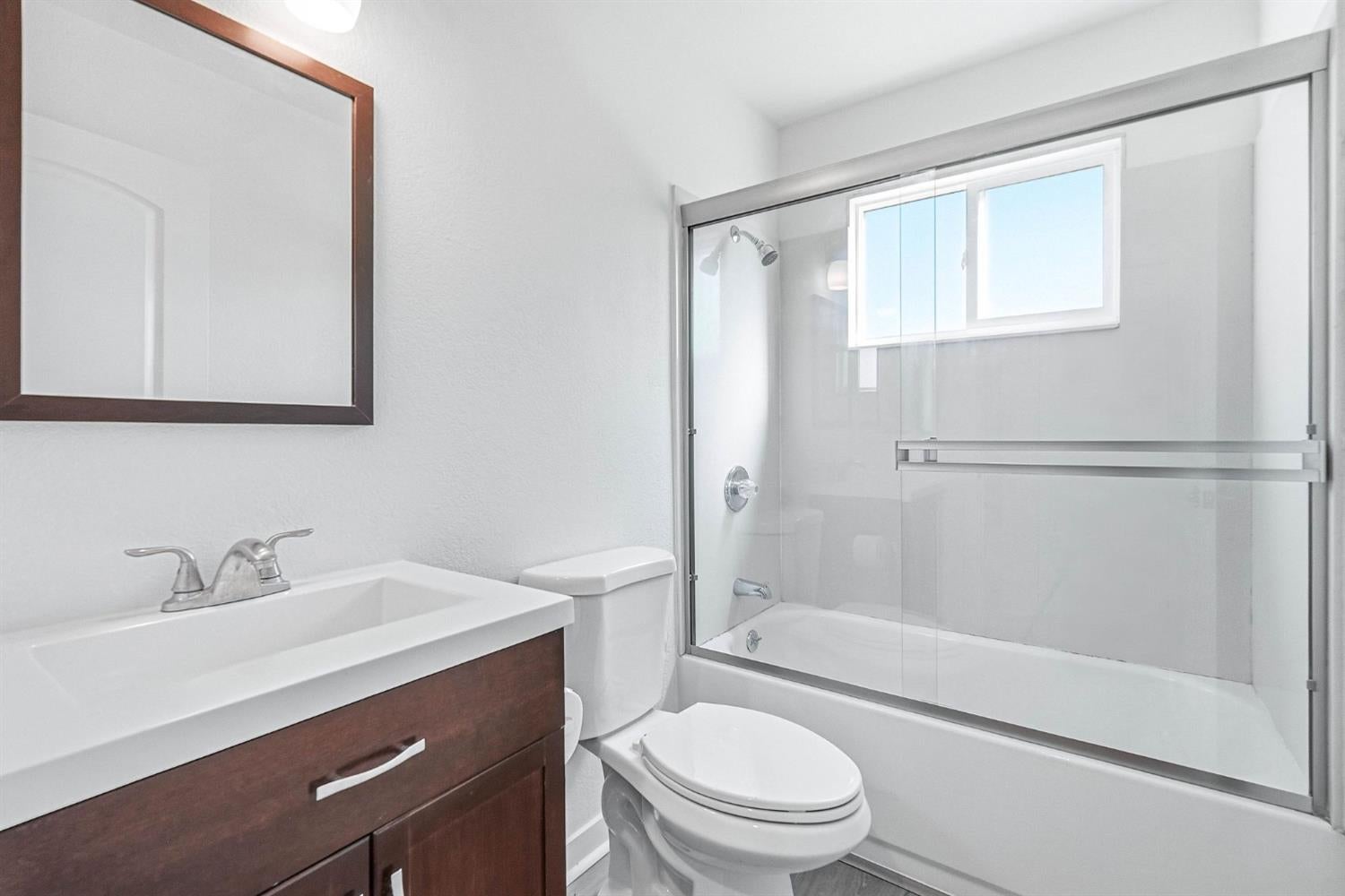 Bathroom with dark wood vanity, toilet, and glass-enclosed tub beneath a high window.