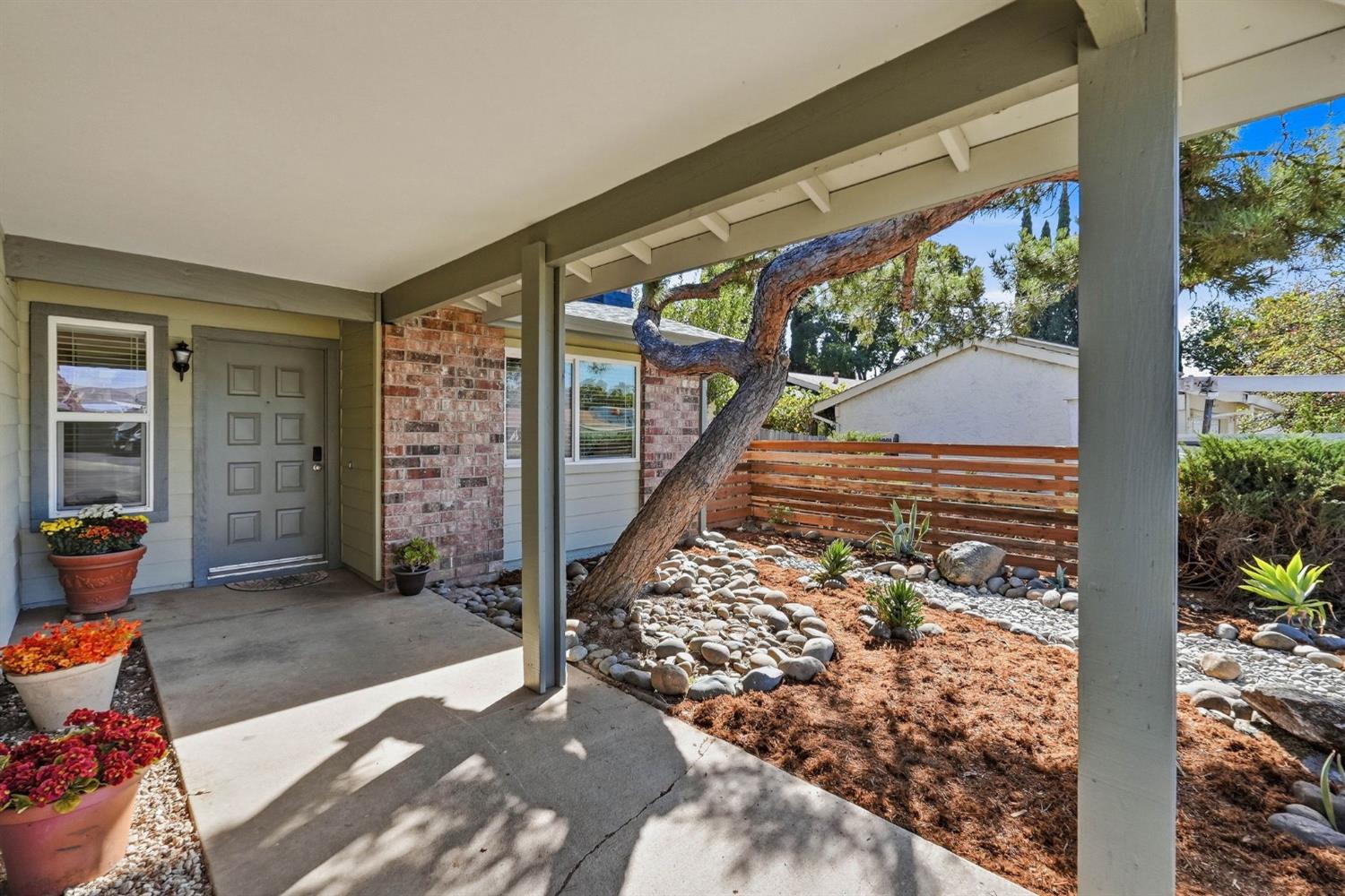 Covered home entry with brick facade, potted flowers, and a rock garden beneath a mature tree.