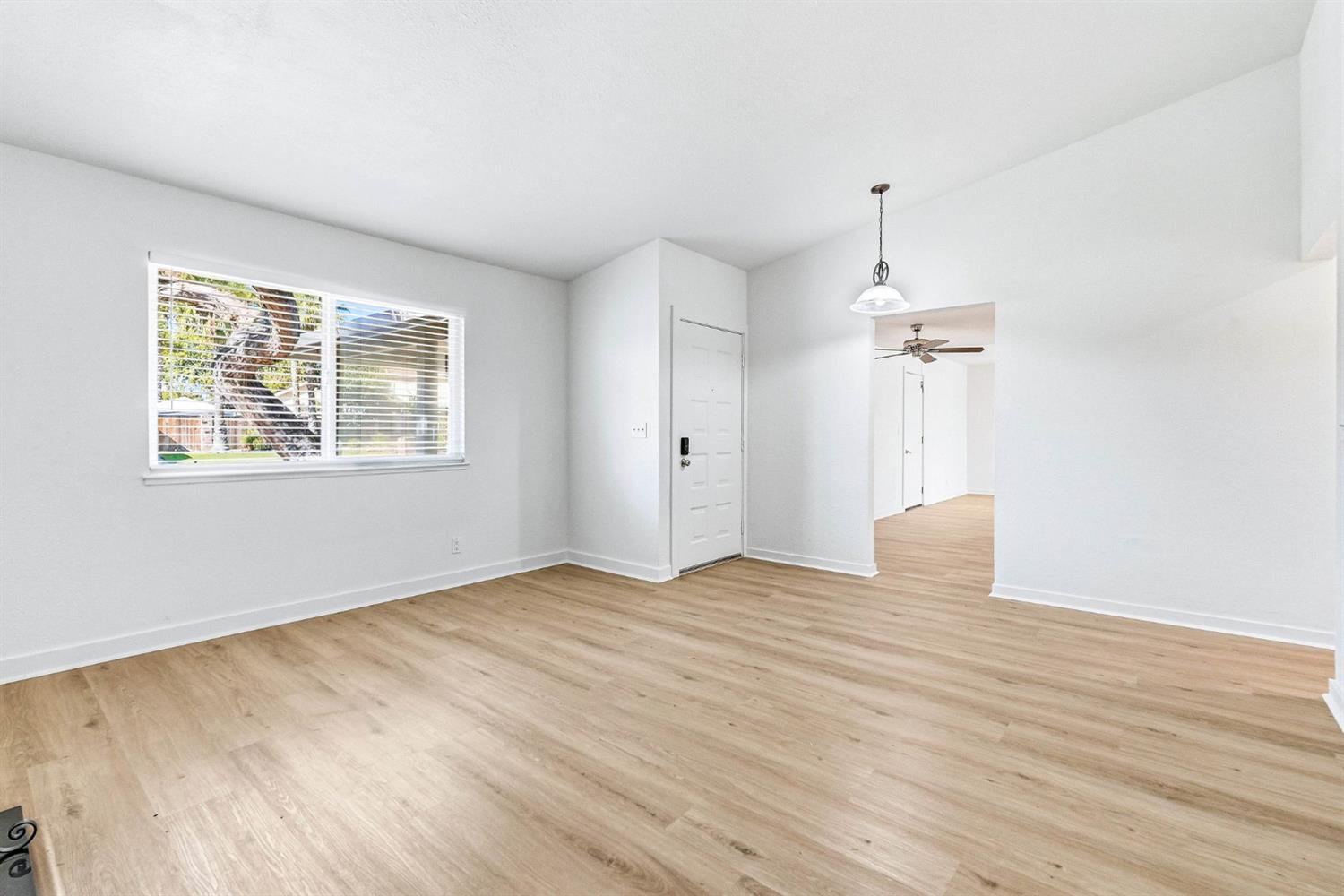 Vacant living room with light wood-look floors, a wide window, and an open hallway.