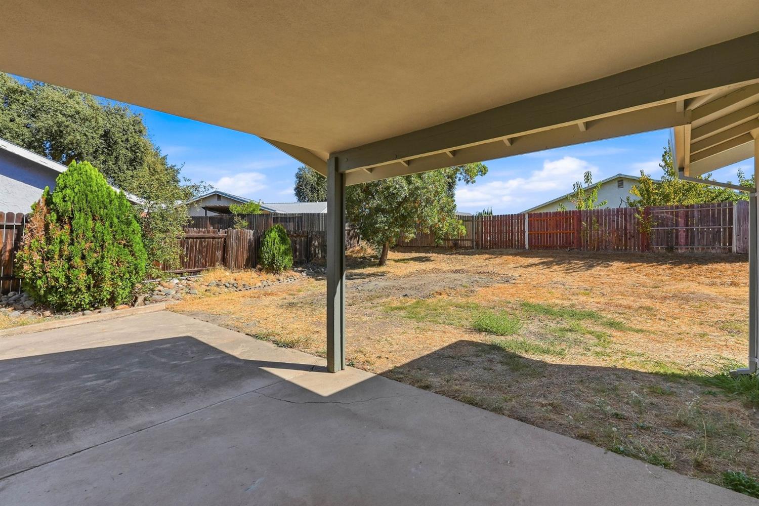 Covered patio overlooking a fenced backyard with dry grass and mature trees.