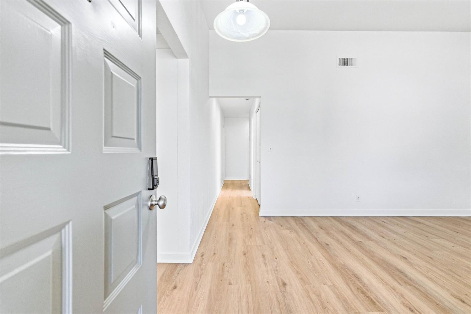 Empty room with light wood-look flooring and a hallway beyond a partially open entry door.