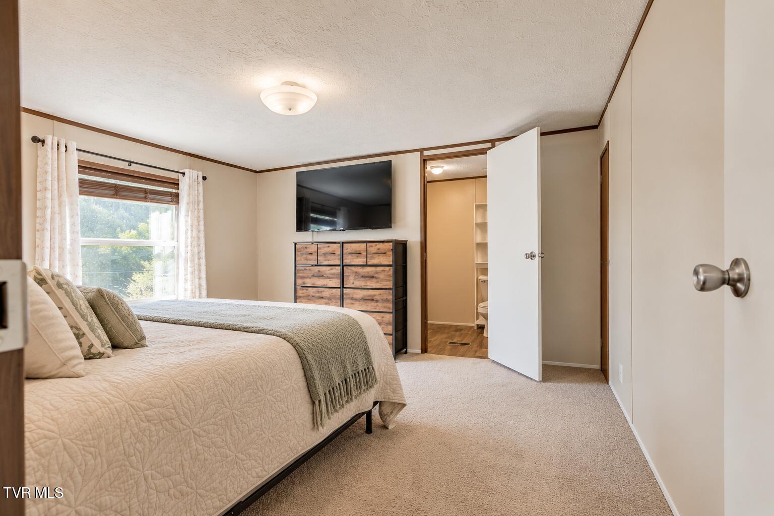 Bedroom with a large quilted bed, wood-finish dresser, television, and open doorway to a bathroom.