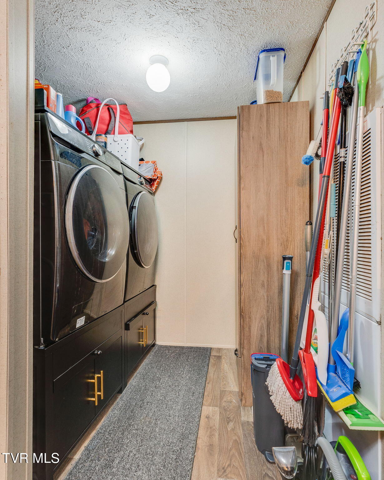 Laundry room with front-loading washer and dryer, dark cabinetry, and cleaning tools along the wall.
