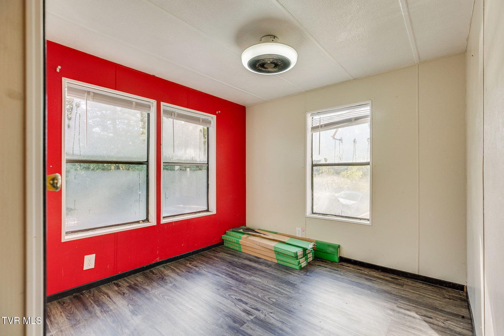 Empty room with a red accent wall, three windows, and dark wood-look flooring.