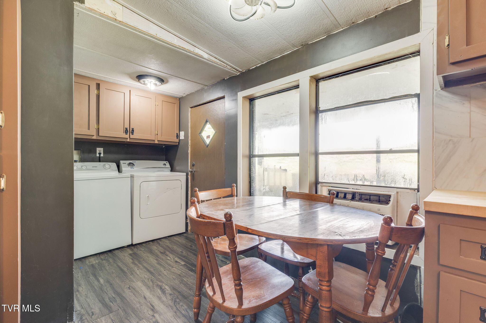 Laundry room with a washer and dryer beside a wooden dining table and large windows.