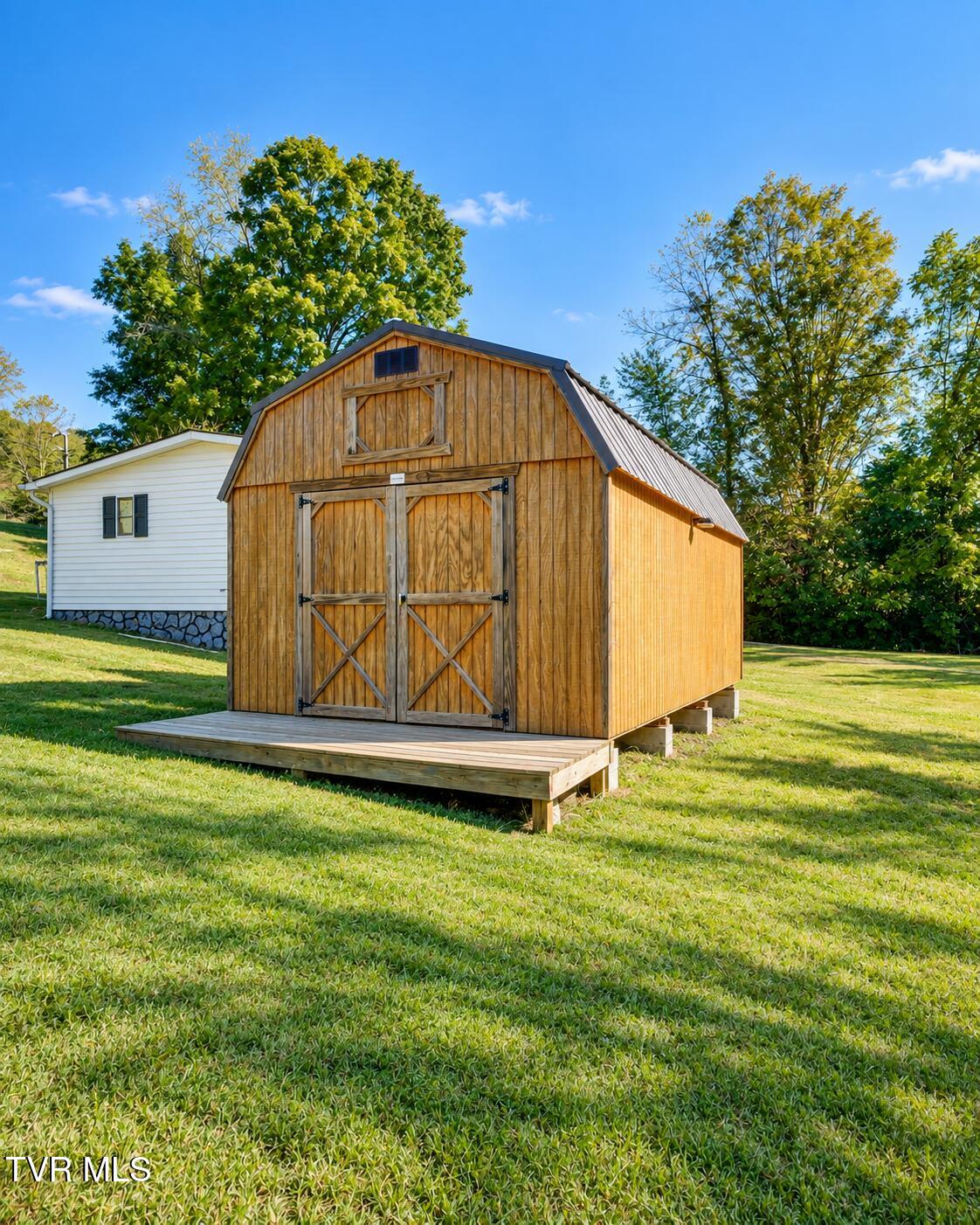Wooden gambrel-roof shed with barn doors and a raised deck on a grassy lawn.