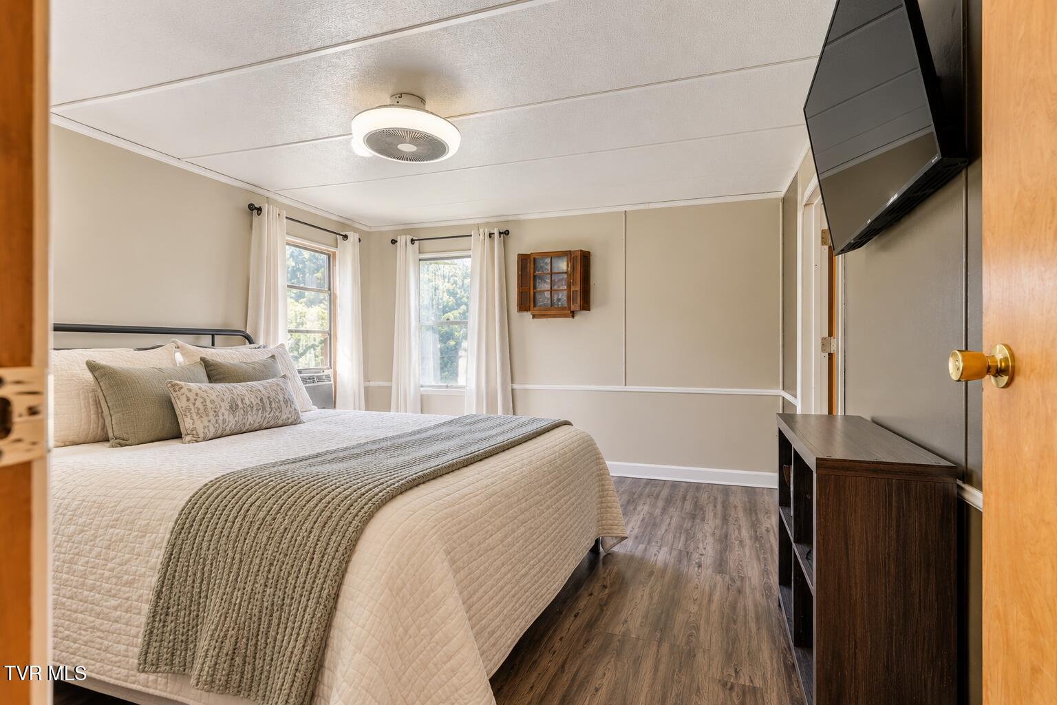 Bedroom with a cream quilted bed, gray throw, two windows, and a wall-mounted television.