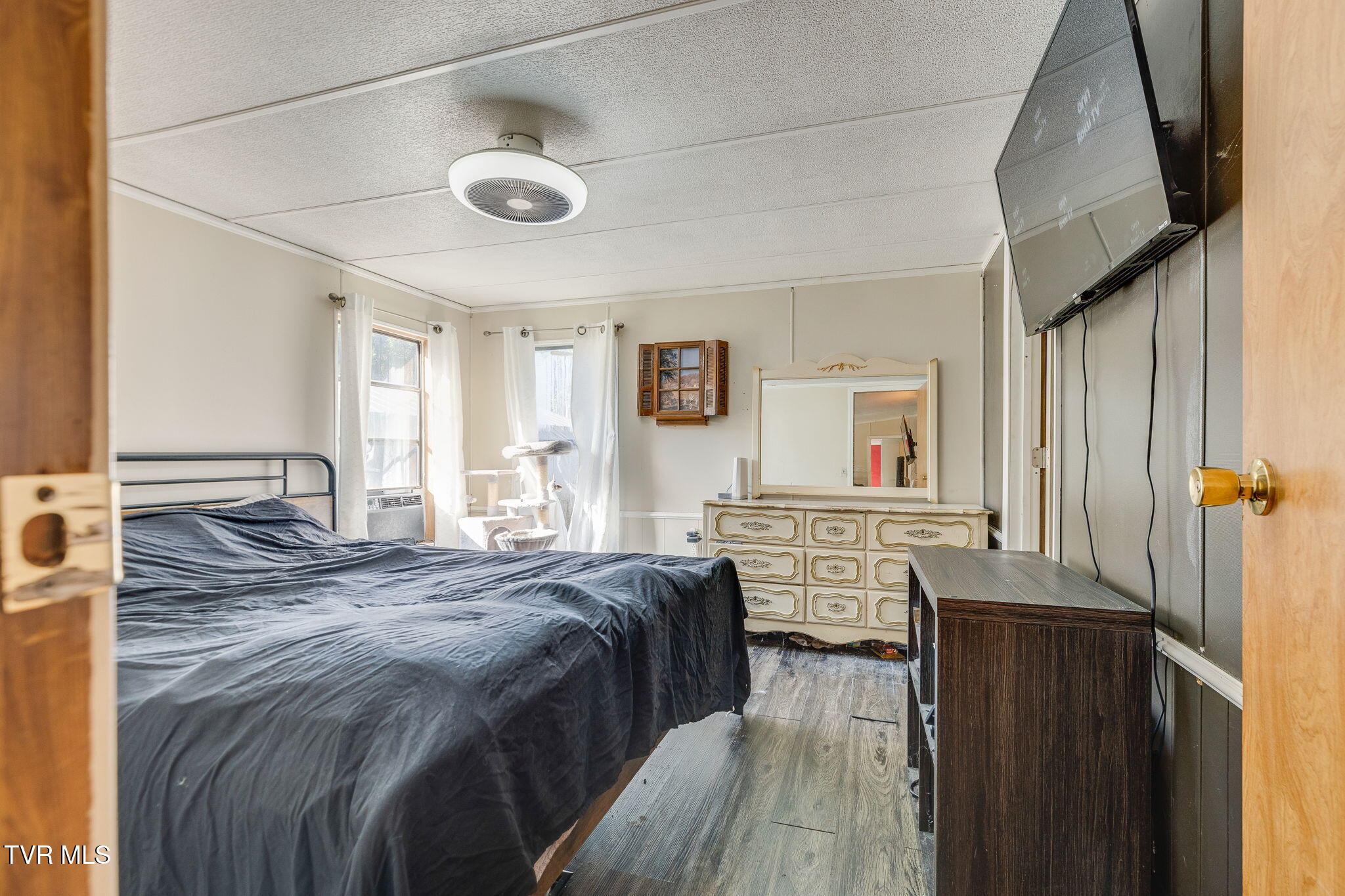 Bedroom with dark bedding, ornate cream dresser and mirror, and a wall-mounted television.