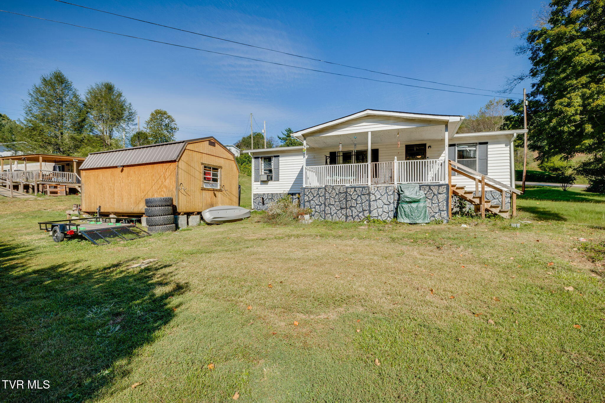 White single-story home with a covered porch and stone foundation, beside a wooden shed and trailer.