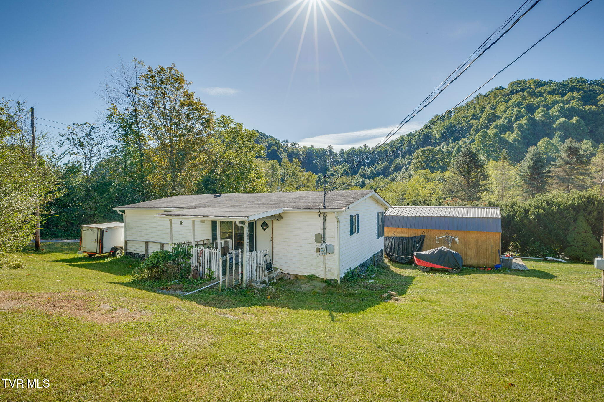 White single-story house with a fenced patio and metal-sided outbuilding, backed by wooded hills.