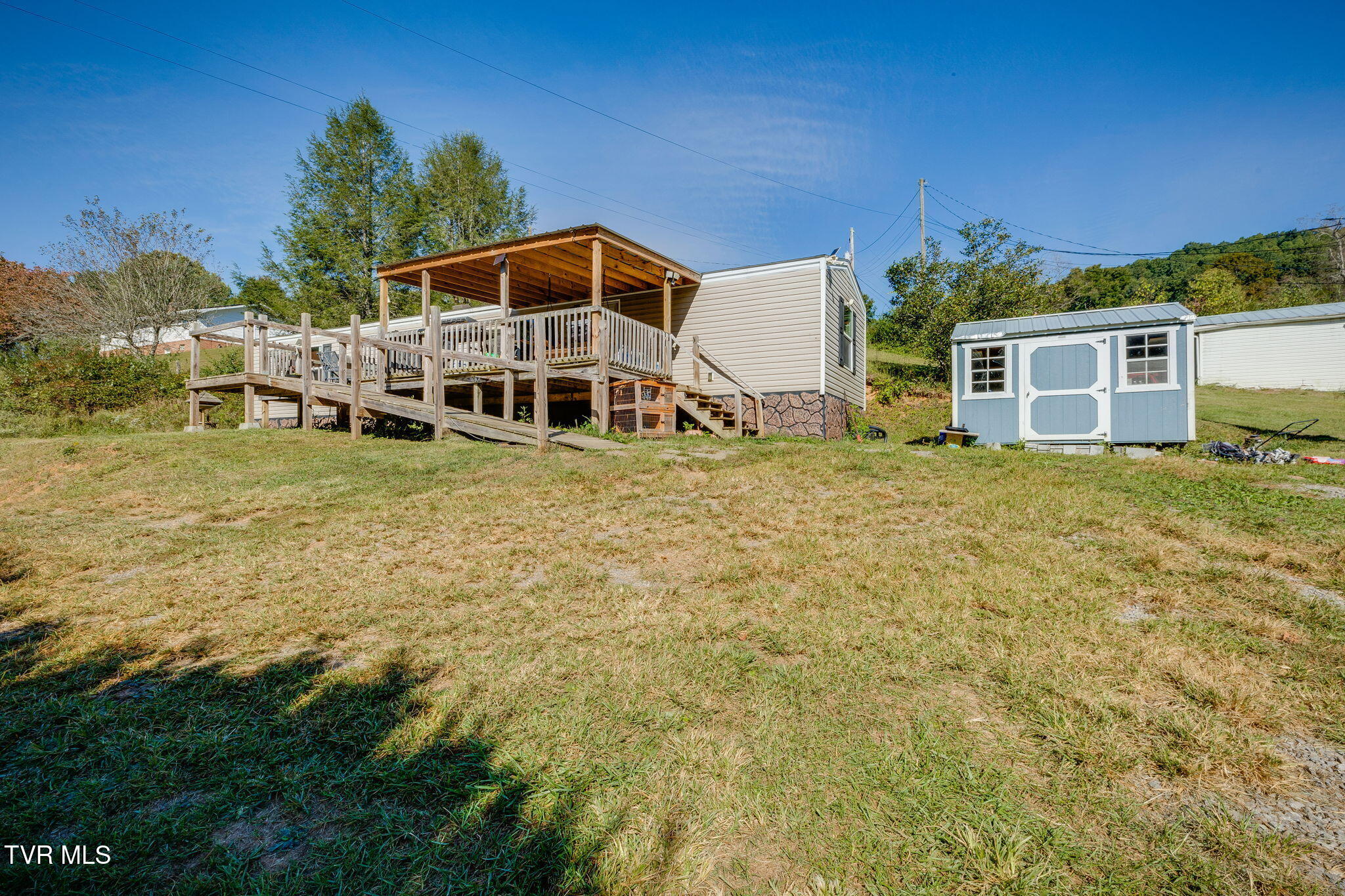 Manufactured home with a large covered wooden deck and blue-and-white storage shed on a grassy slope.