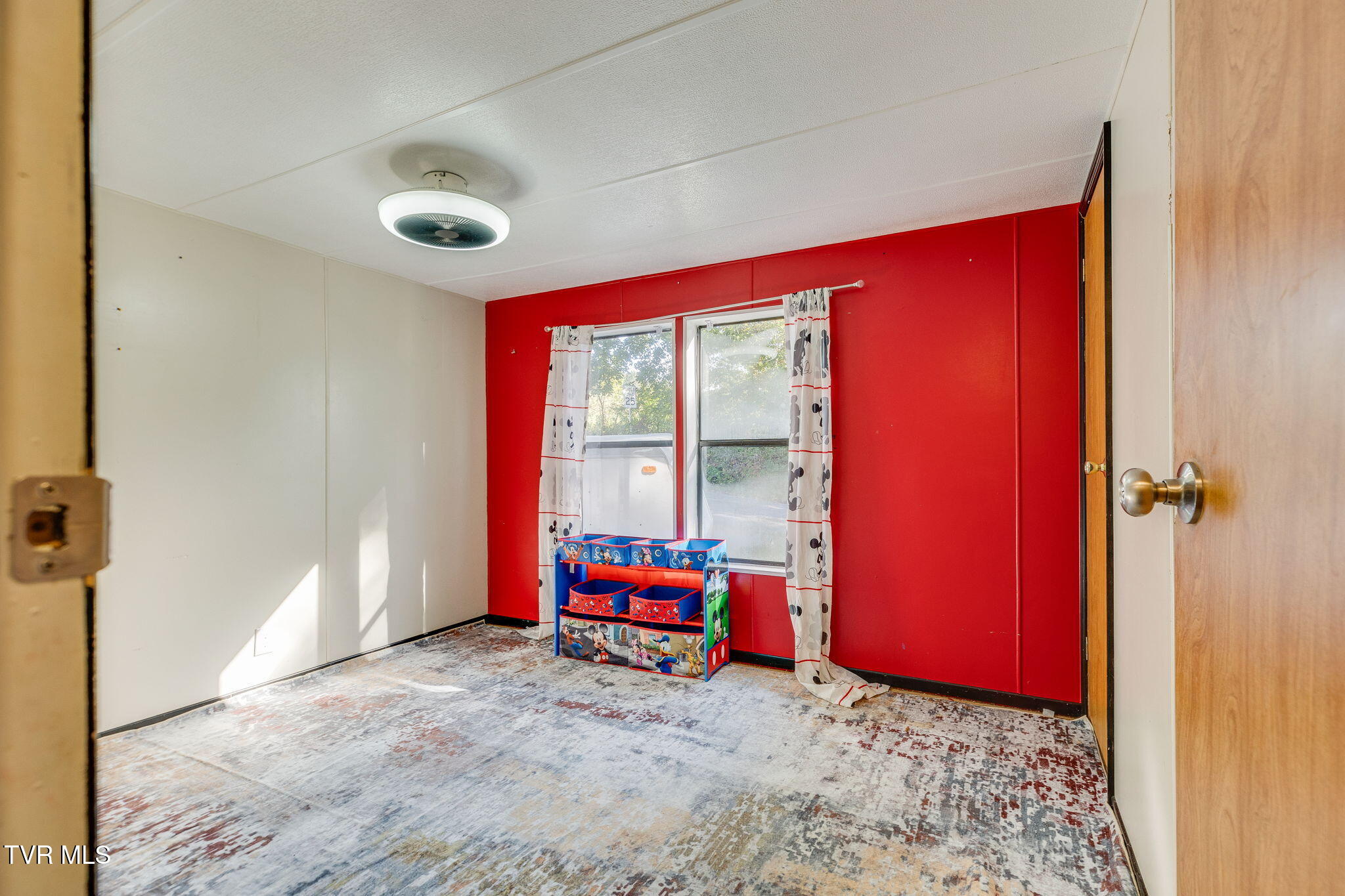 Bedroom with a red accent wall, two curtained windows, and a colorful toy shelf