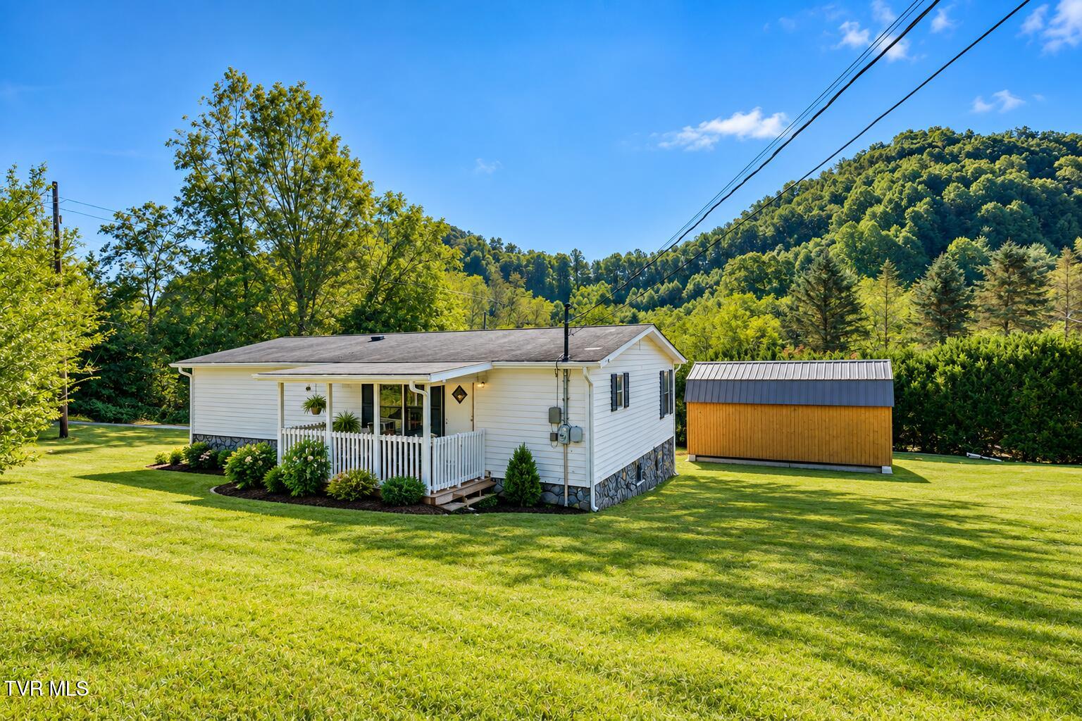 White single-story home with a covered porch and detached wooden shed on a lawn backed by wooded hills.