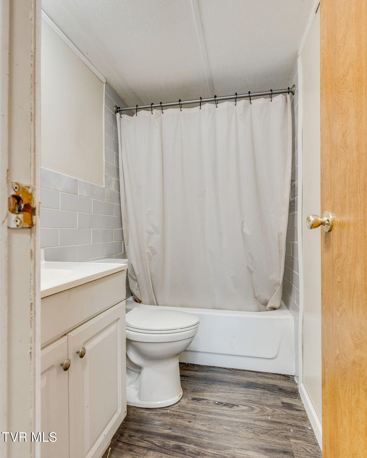 Bathroom with a curtained tub, toilet, cream vanity, gray tile, and dark wood-look flooring.