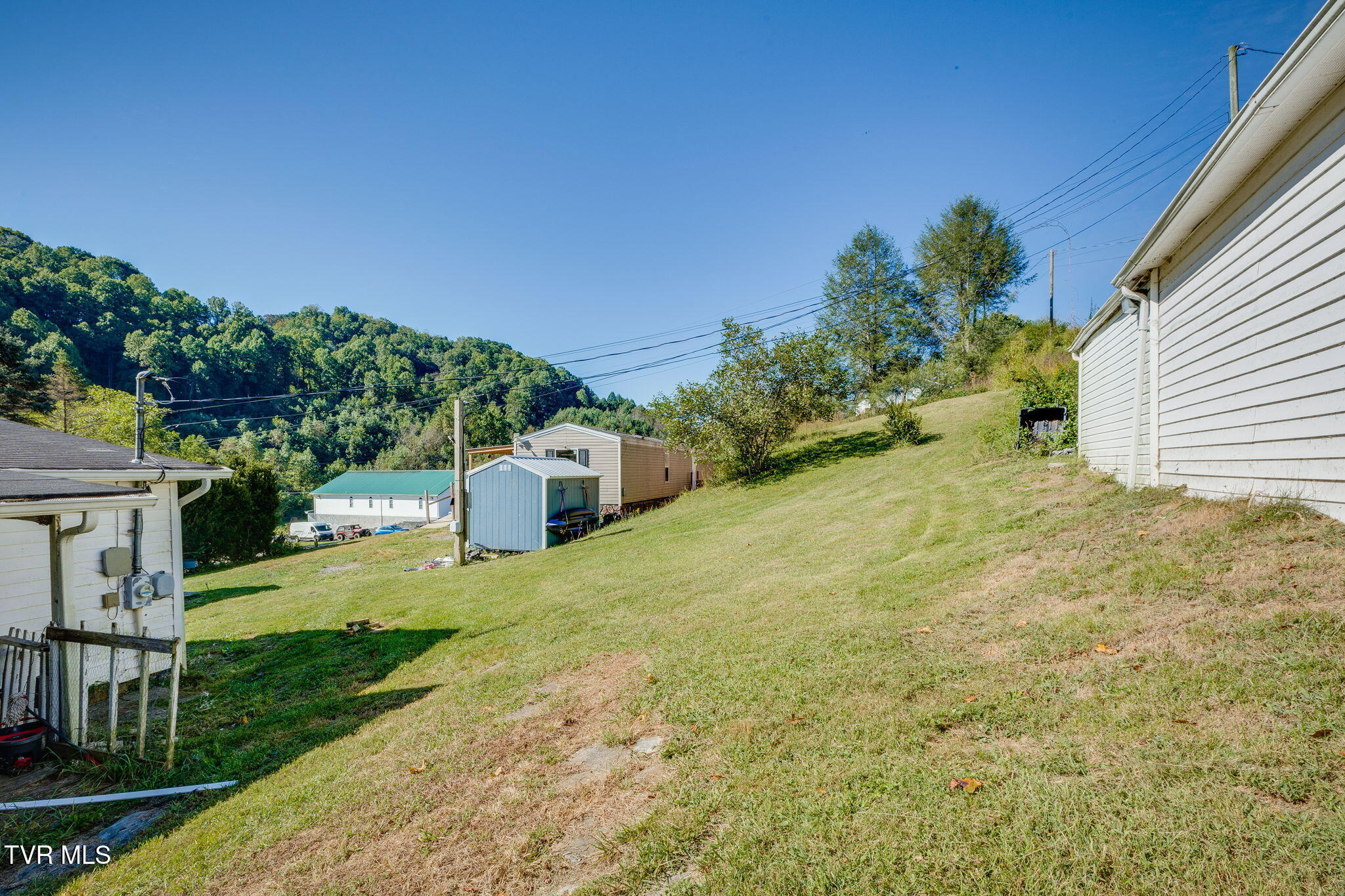 Sloping grassy yard with sheds, neighboring buildings, and a wooded hillside.