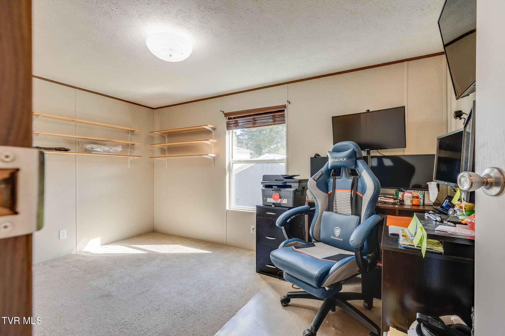 Home office with wall shelves, a cluttered computer desk, and a blue gaming chair.