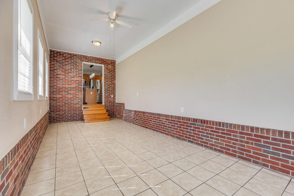 Empty tiled room with brick wainscoting and tall windows, opening to a kitchen.
