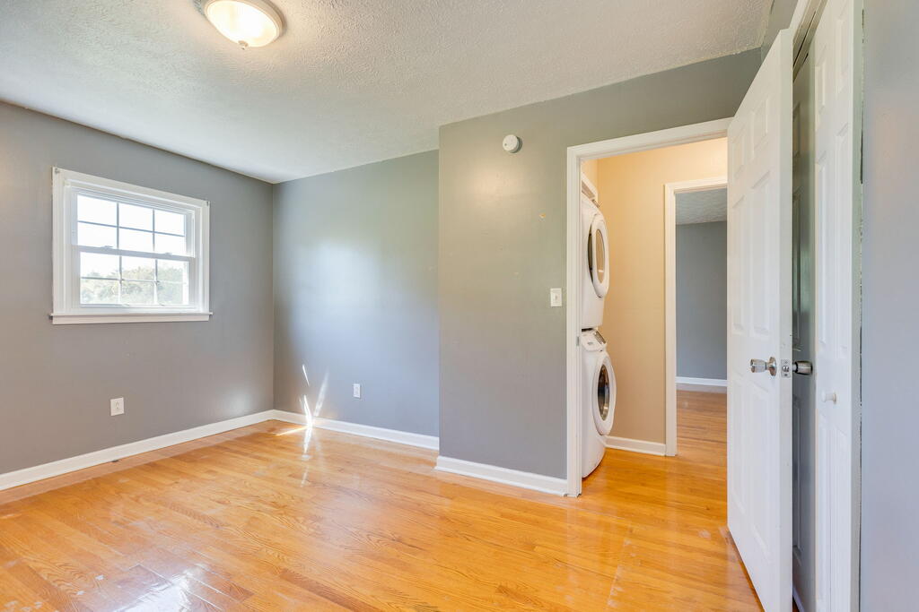 Empty gray room with hardwood floors and a window, opening to a laundry area with stacked washer and dryer.