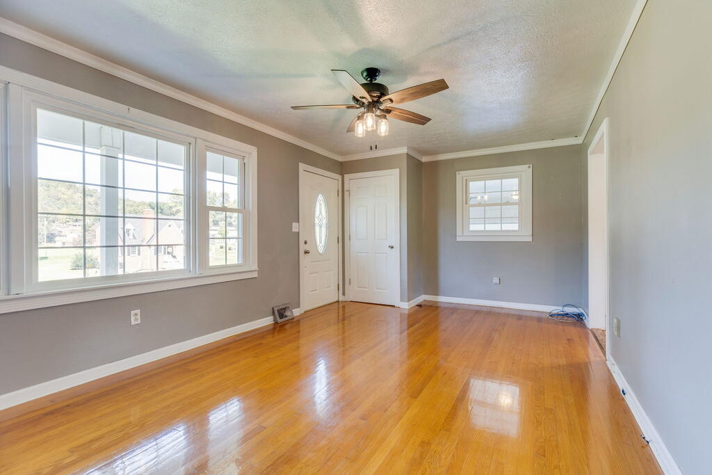 Empty living room with hardwood floors, a ceiling fan, and large front windows.