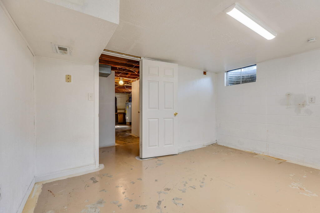 Vacant basement room with white walls, a worn concrete floor, and an open door to a utility area.