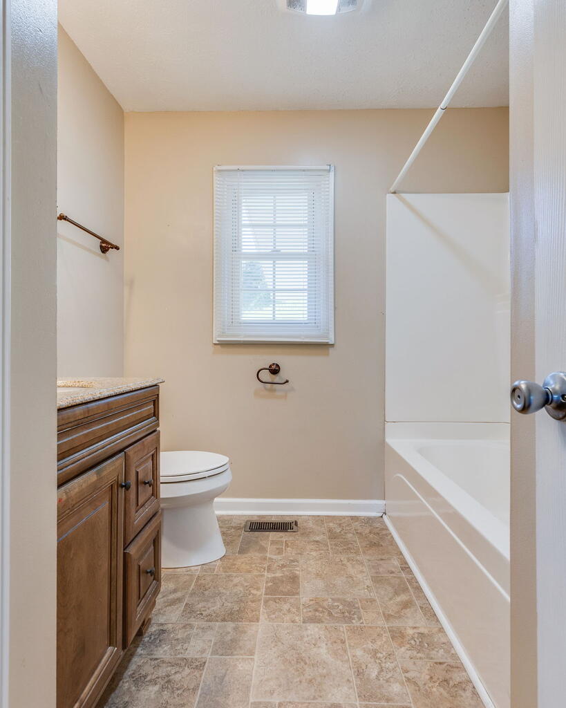 Bathroom with a wood vanity, toilet, tub-shower, and window with blinds.