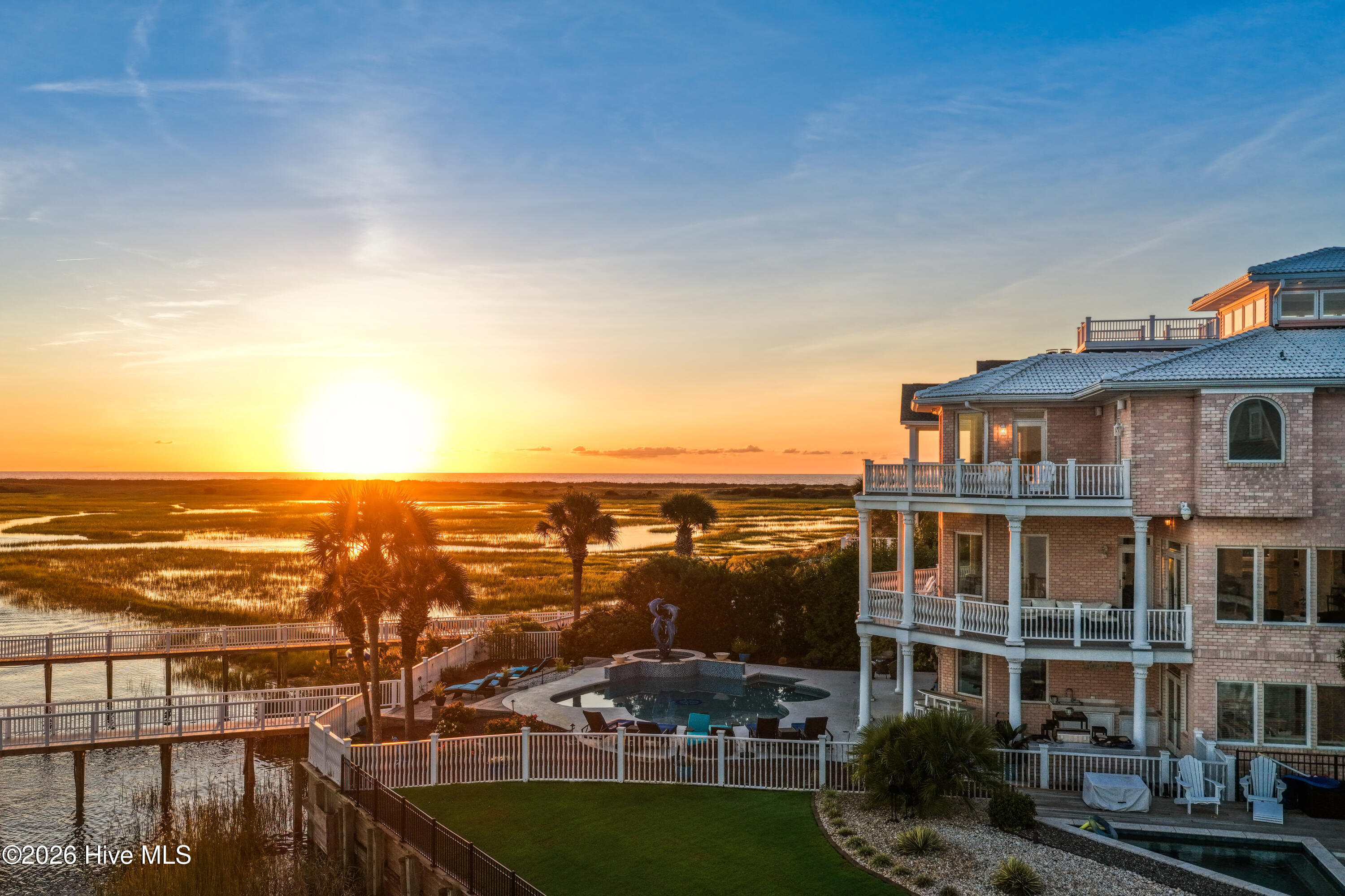 Coastal brick home beside a pool, with marsh and ocean views at sunset.