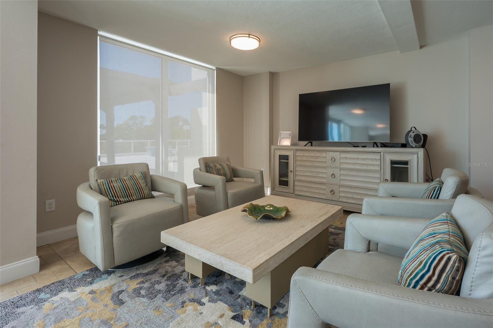 Condo living room with pale seating, striped pillows, a stone coffee table, and a television by broad windows.