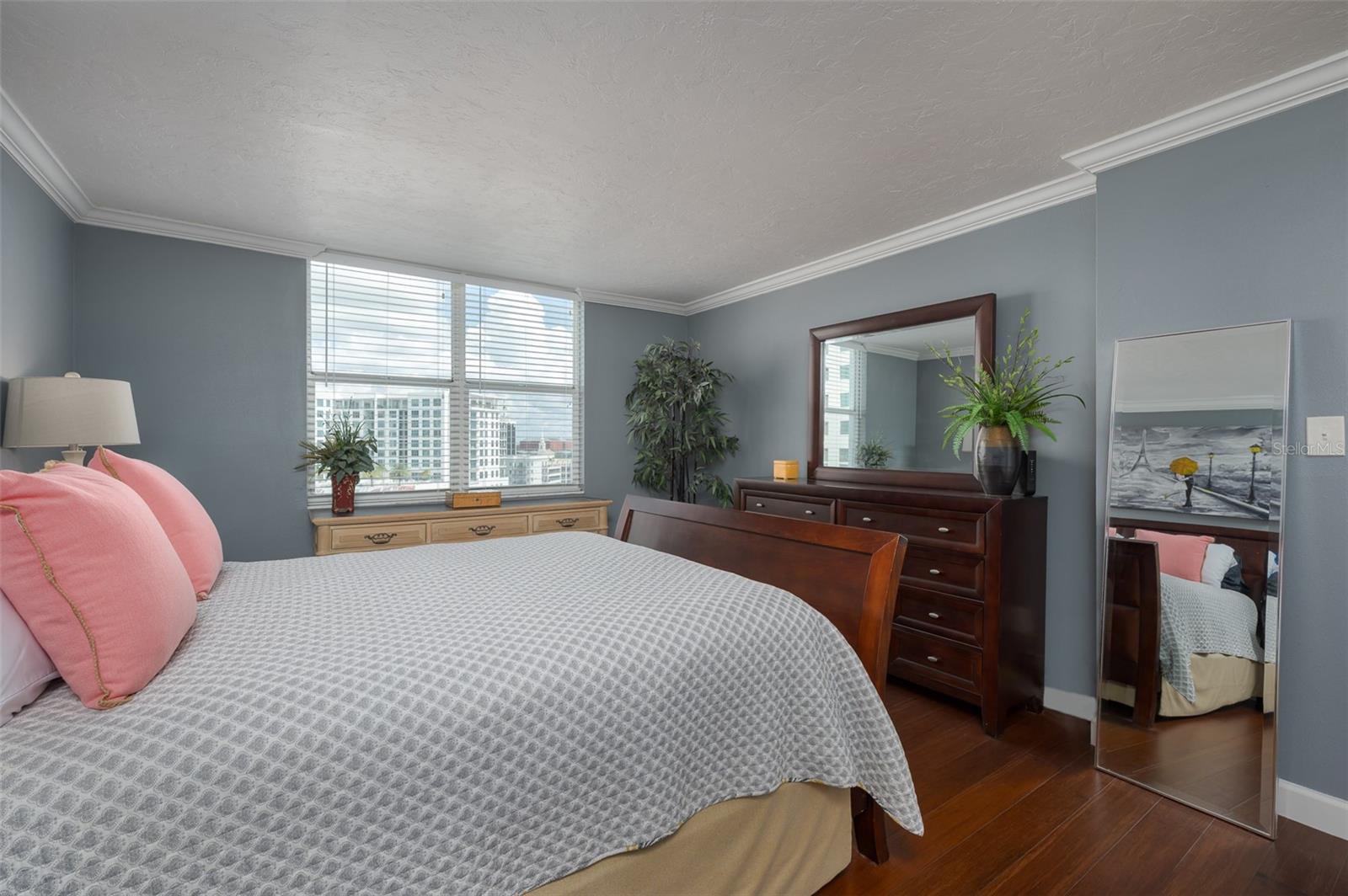 Bedroom with pink pillows, dark wood furniture, and windows overlooking nearby buildings.