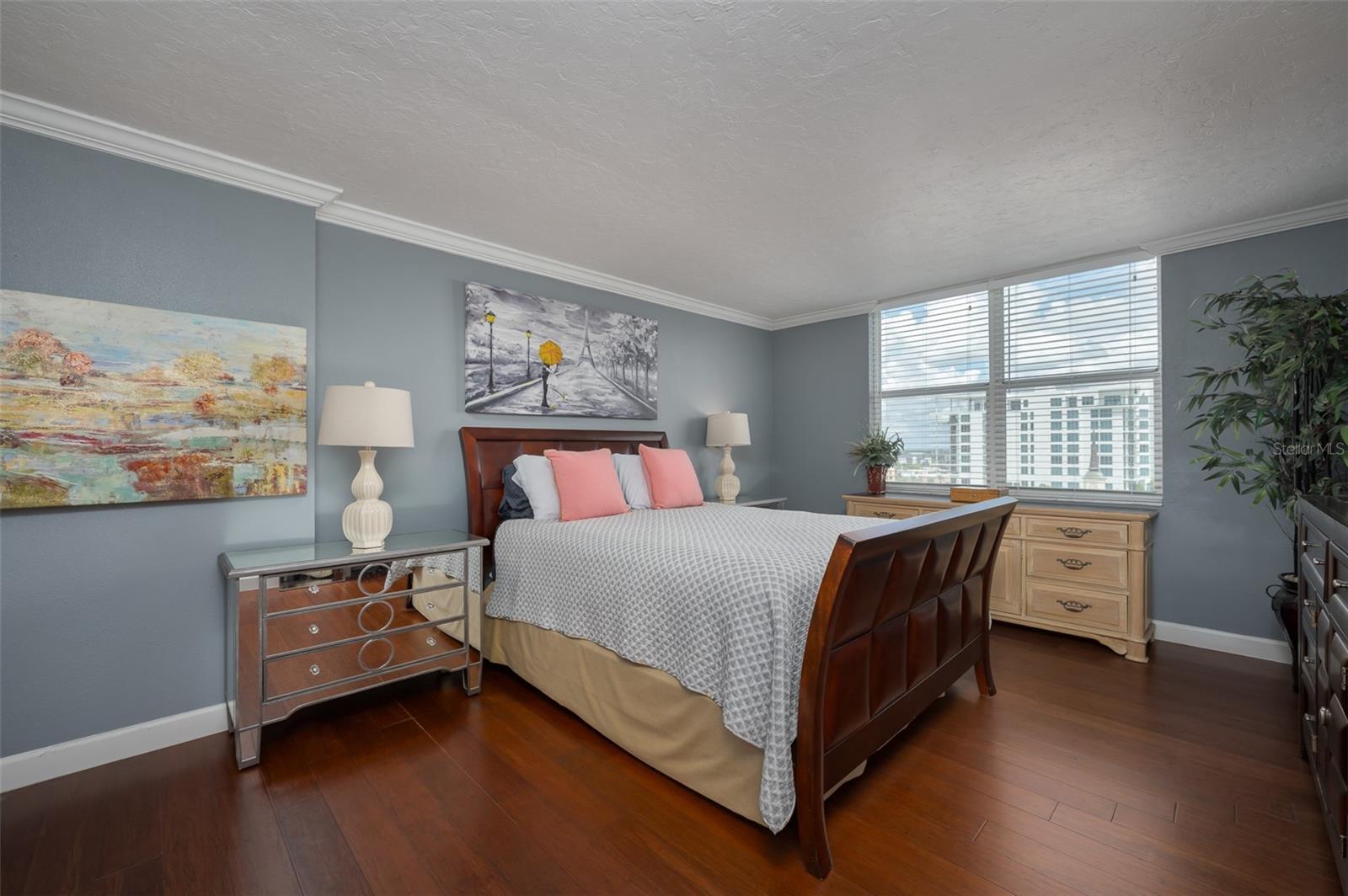 Furnished bedroom with a dark wood bed, coral pillows, and large windows overlooking nearby buildings.