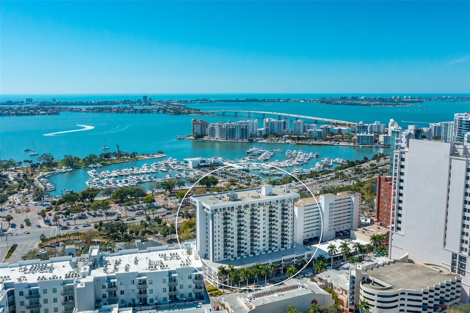 Waterfront condominium highlighted by a circle, overlooking a marina, bay, and distant skyline.