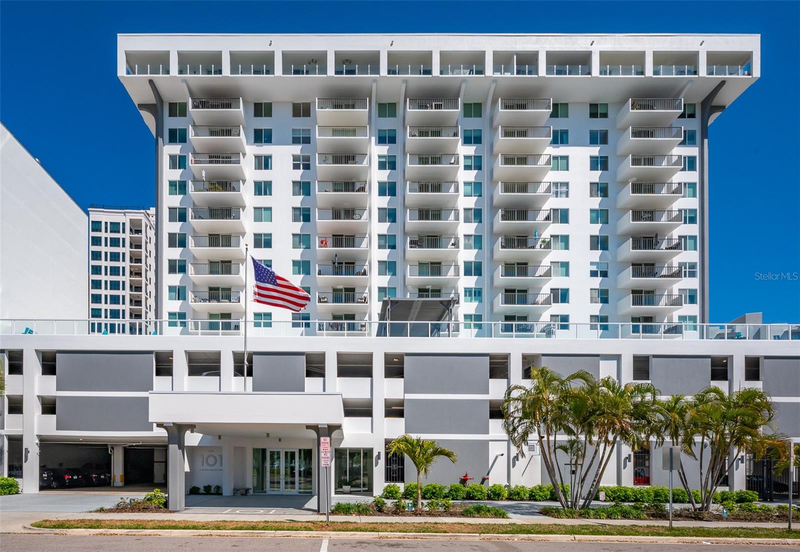 White condominium tower with stacked balconies, a covered entrance, palm trees, and an American flag.