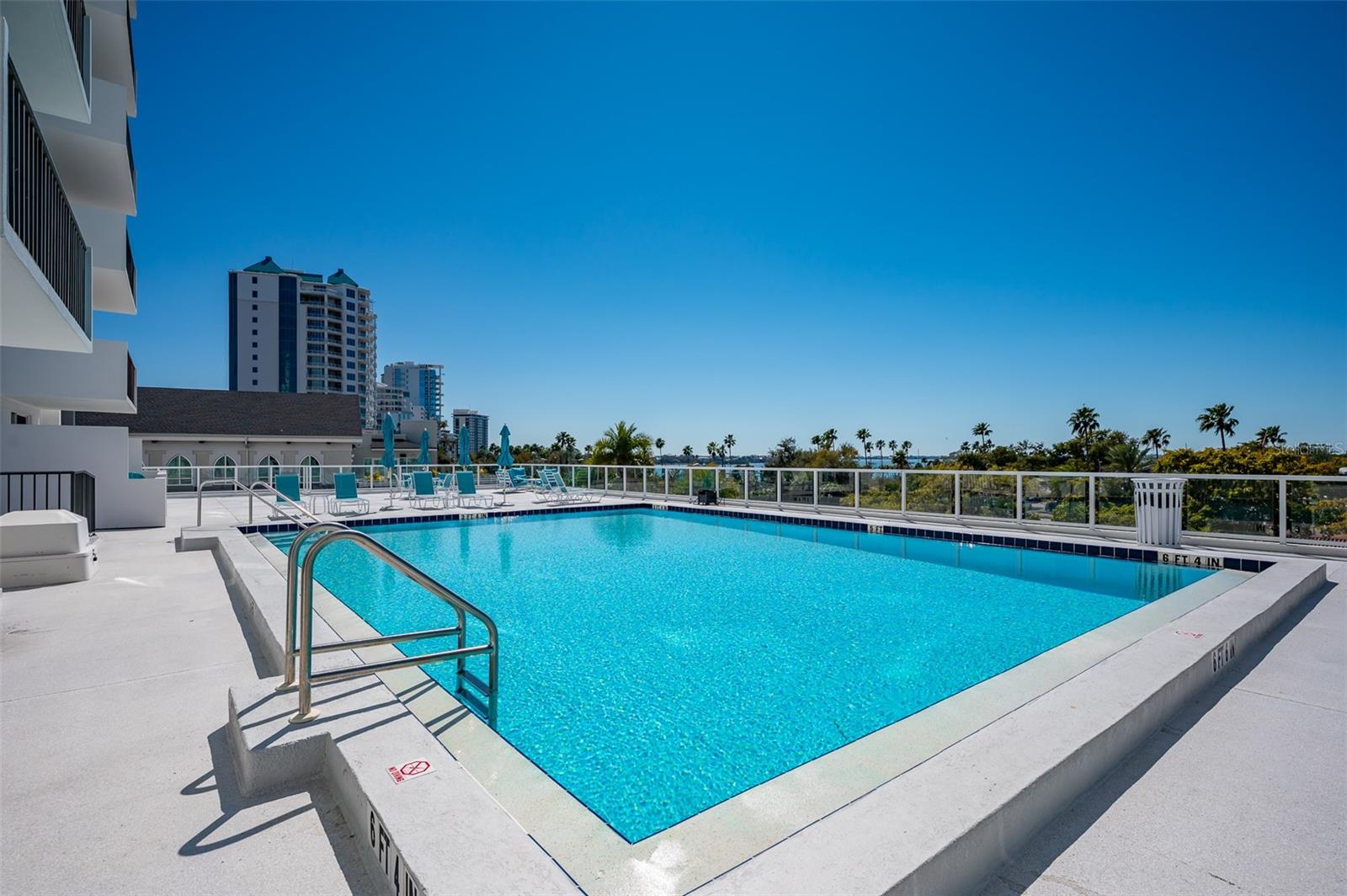 Rooftop condominium pool with a metal ladder, lounge chairs, and palm-lined surroundings.