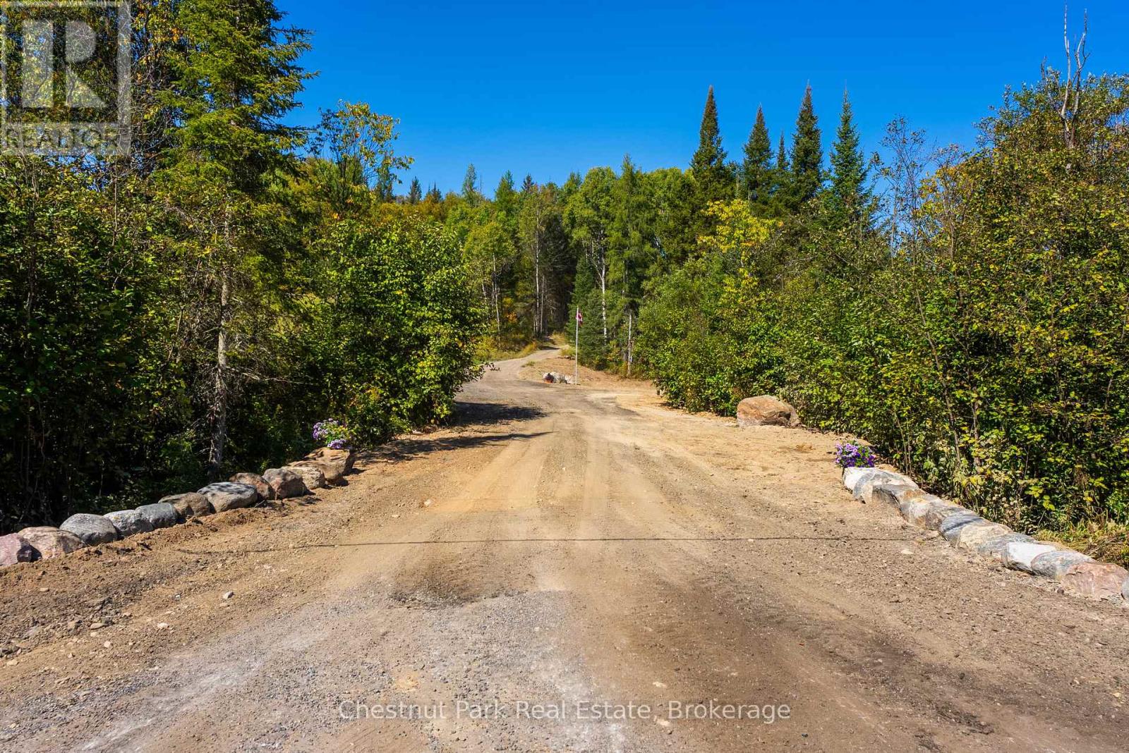 Gravel entrance road lined with rocks and flowers, leading into wooded property.