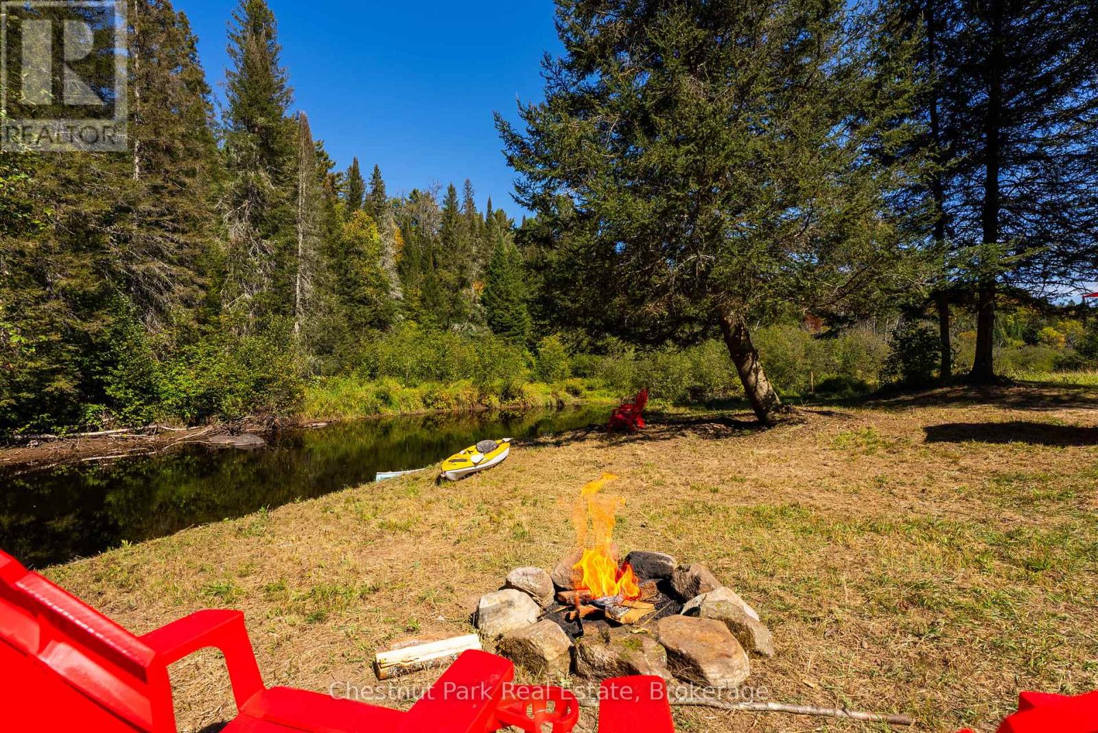 Wooded waterfront clearing with a stream, yellow kayak, red Adirondack chairs, and a stone fire pit.