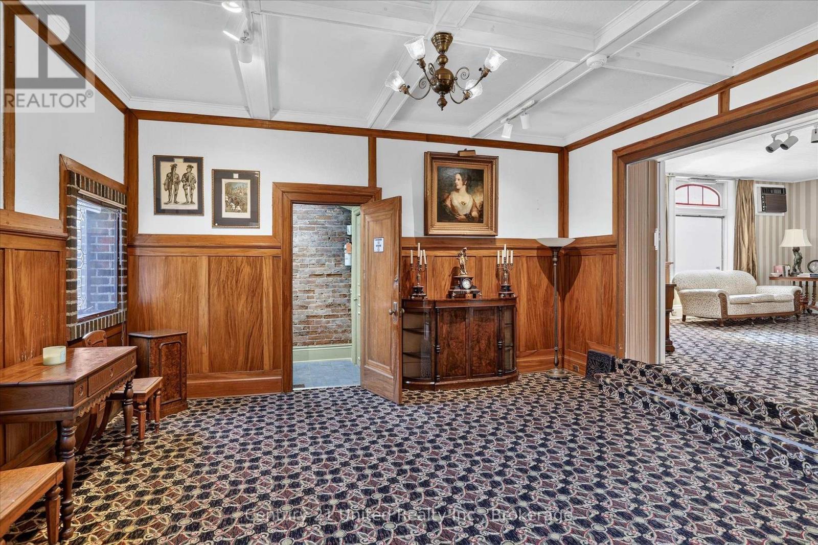 Historic room with wood-paneled walls, patterned carpet, antique furnishings, and an open doorway to a sitting room.