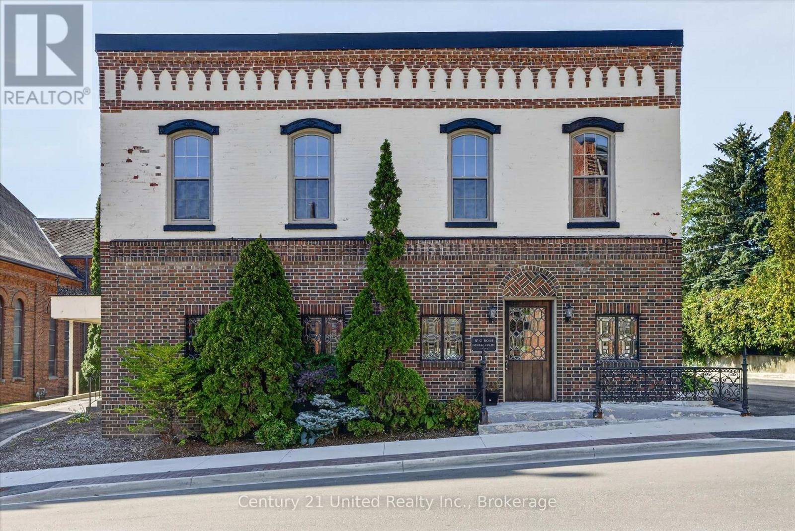 Two-story historic brick building with a white upper facade, decorative brickwork, and landscaped front entrance.