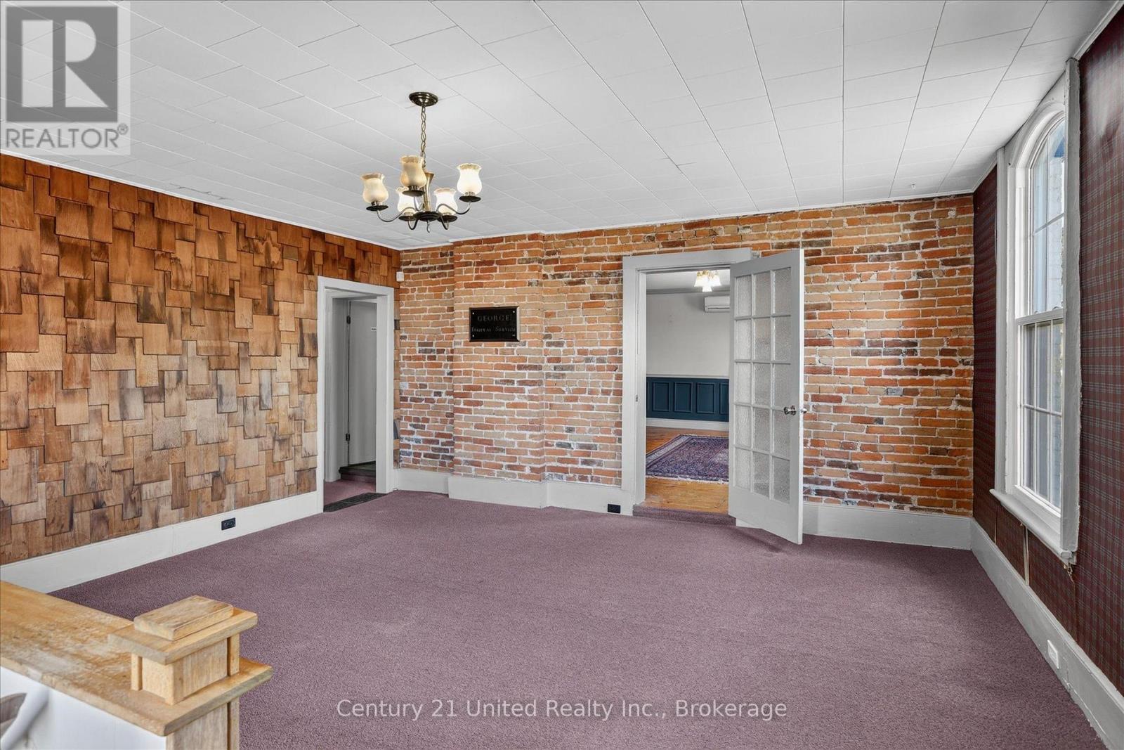 Spacious brick-walled room with mauve carpet, a chandelier, wood feature wall, and tall arched window.