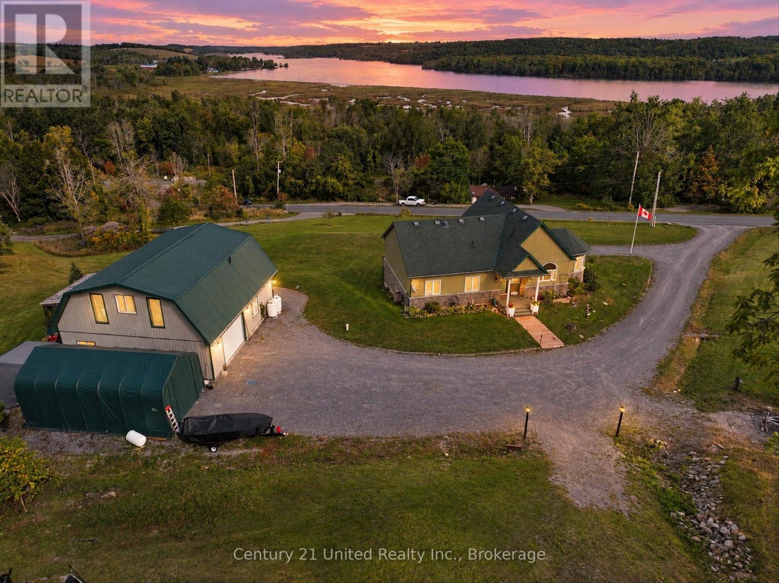 Rural home and outbuilding beside a gravel drive, with a lake and forest beyond.