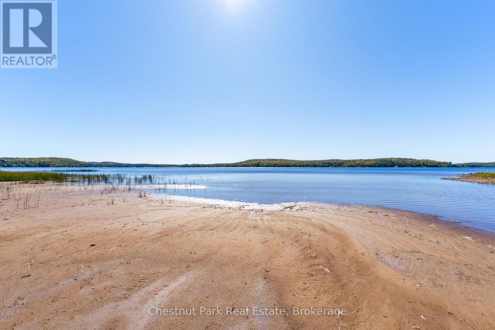 Sandy beach curves into a calm lake, with reeds at the water’s edge and wooded hills across the water.