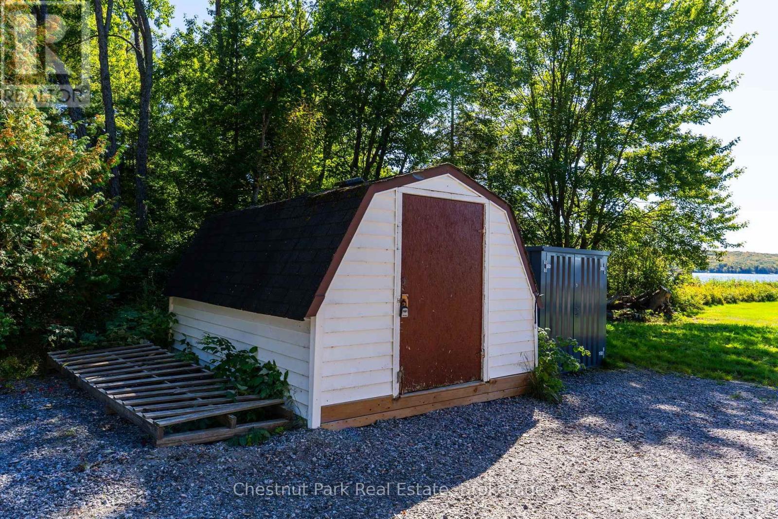 White storage shed with a dark gambrel roof and reddish-brown door, near a lake.