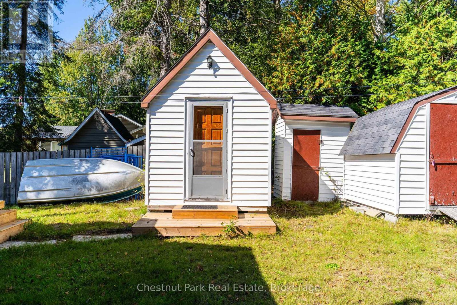 White gable-roofed outbuilding with a wooden entry platform, flanked by sheds and an overturned boat.
