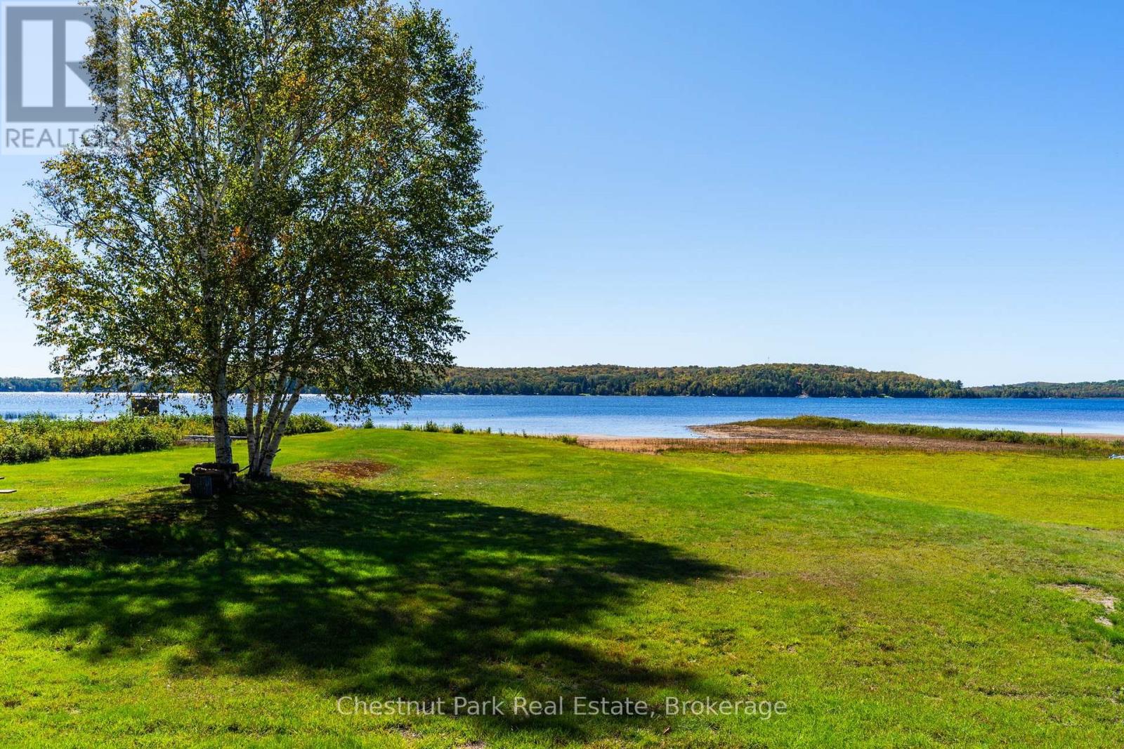 Waterfront lawn beside a lake, shaded by mature trees with a wooded shoreline across the water.