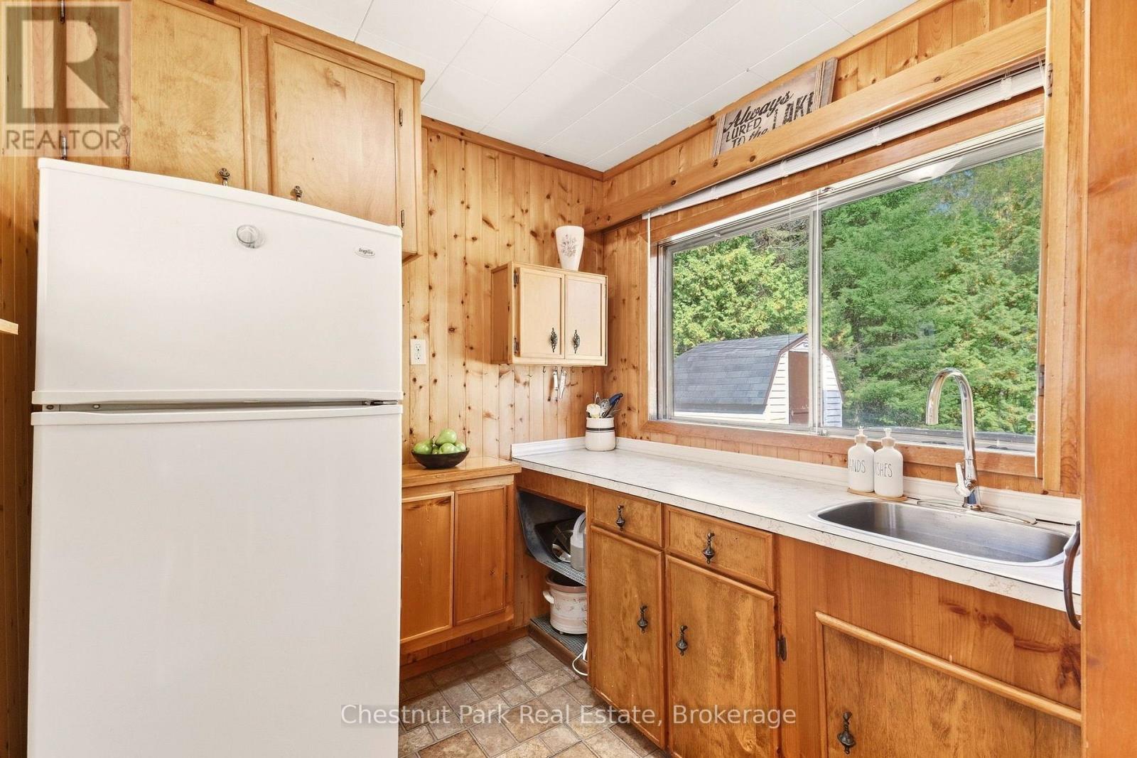 Wood-paneled kitchen with a white refrigerator, long counter, double sink, and wooded view through the window.