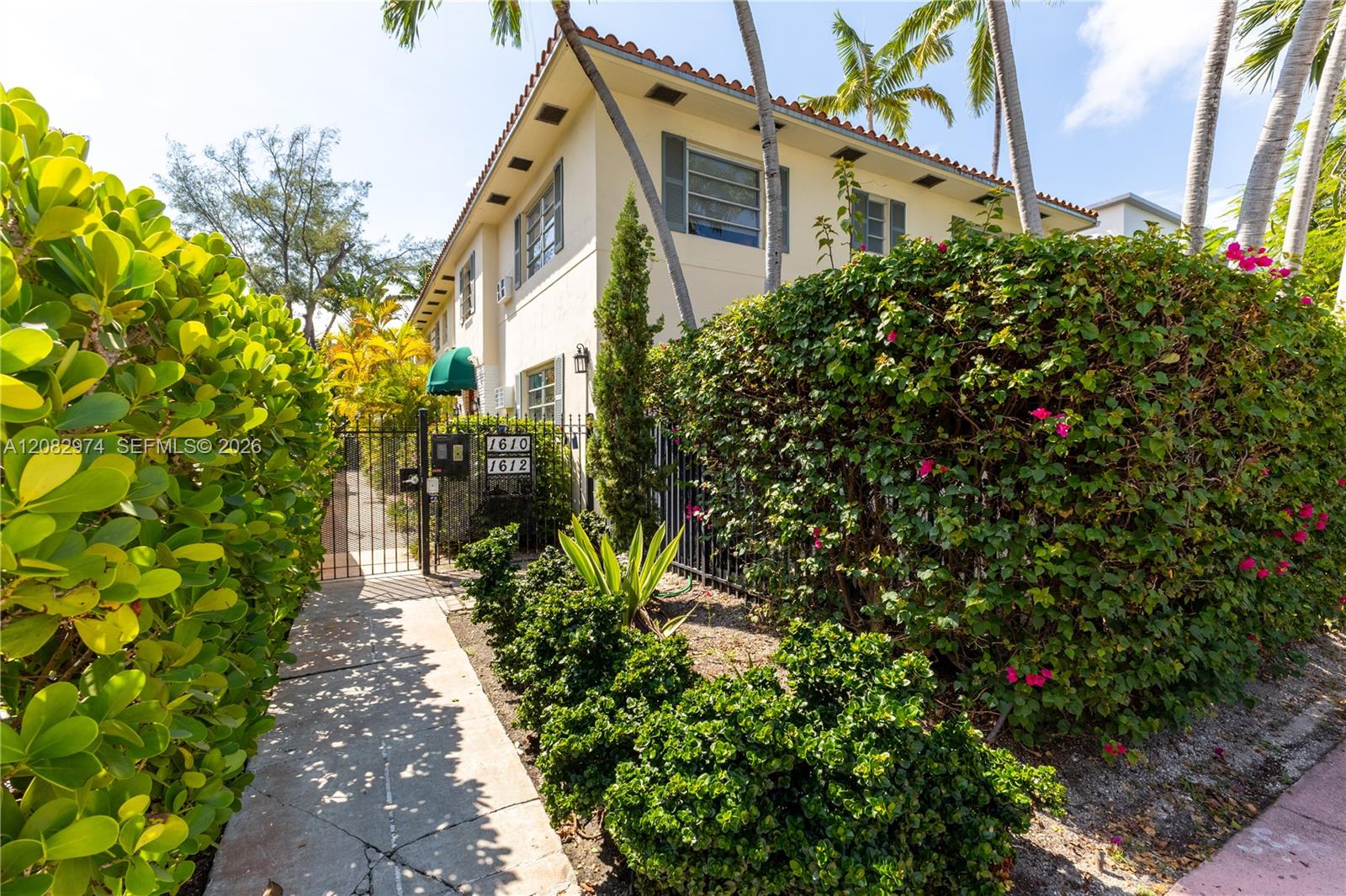 Two-story cream stucco building behind flowering hedges, palms, and a gated walkway.