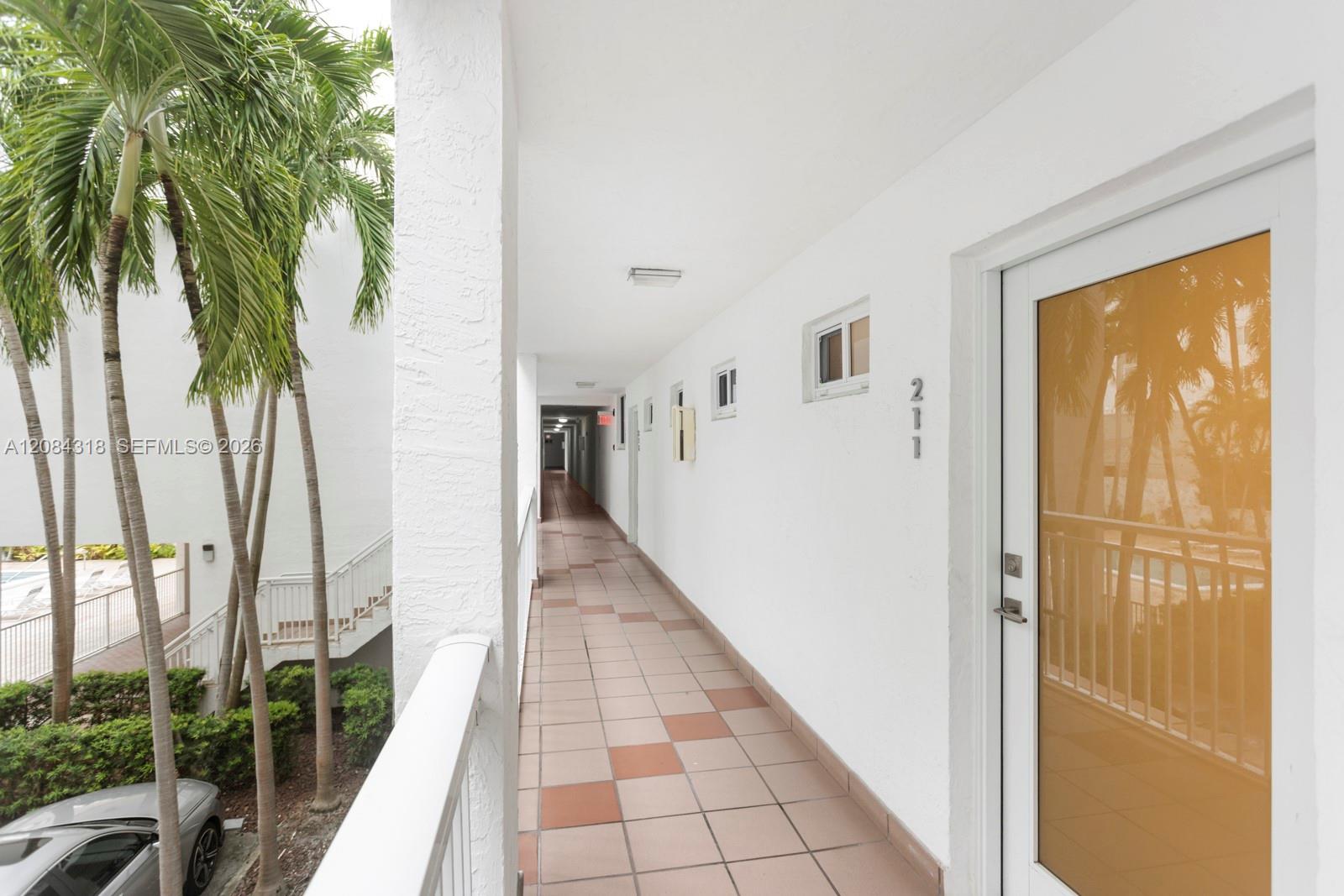 Covered apartment corridor with a tiled floor, glass entry door, and palm trees outside.