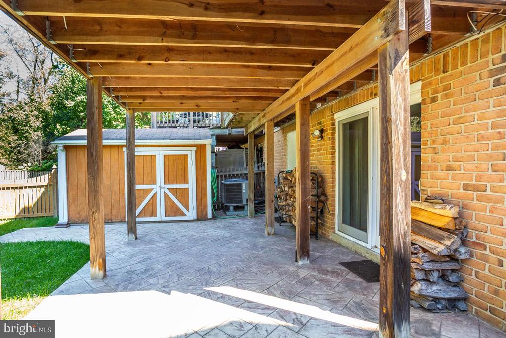 Covered patio with exposed timber beams, stamped concrete, stacked firewood, and a wooden shed.