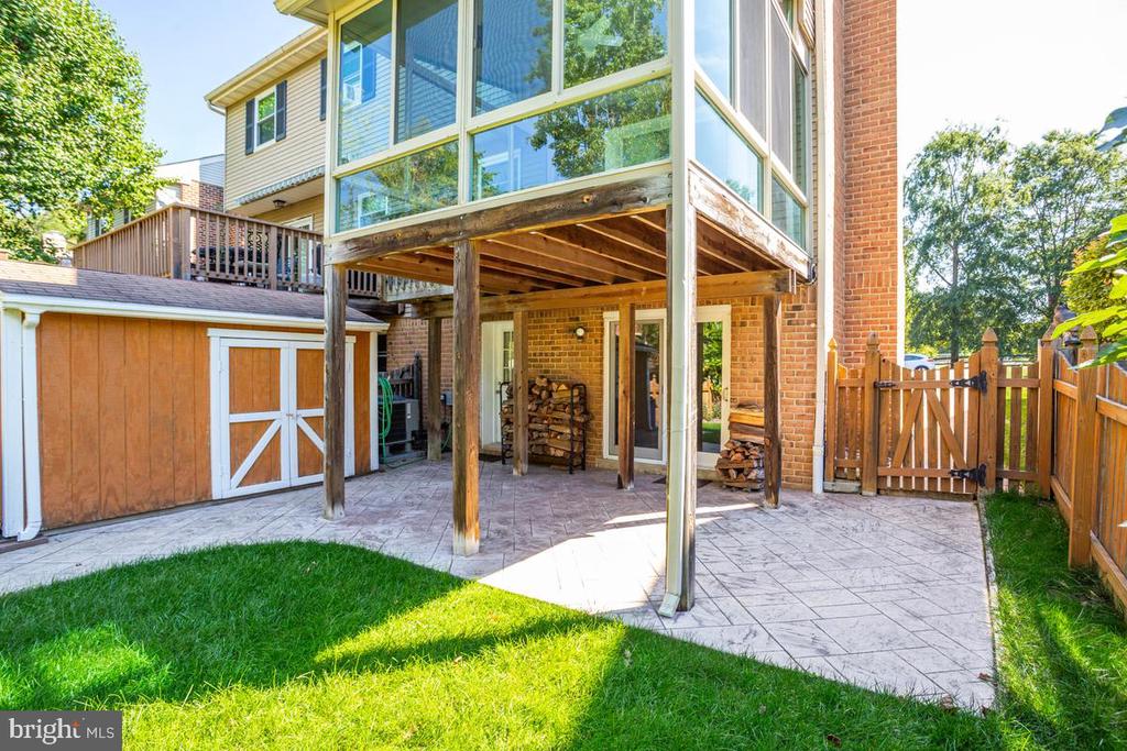 Rear patio beneath a raised deck, beside a wooden shed and fenced lawn.
